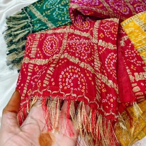 May include: A selection of bandhani scarves in red, green, pink, and yellow. The scarves feature detailed white and gold patterns and gold and red fringing. The text "@jaipursmandecor" is visible on the scarves. The scarves are displayed on a white surface.