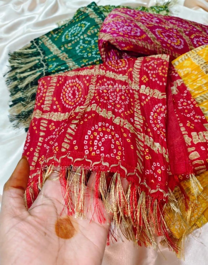 May include: A selection of bandhani scarves in red, green, pink, and yellow. The scarves feature detailed white and gold patterns and gold and red fringing. The text "@jaipursmandecor" is visible on the scarves. The scarves are displayed on a white surface.