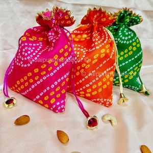 Può includere: Tre colorate sacche con coulisse in rosa, arancione e verde, ciascuna ornata con un motivo bandhani di punti bianchi e gialli. Le sacche sono impreziosite da finiture dorate e cordini, con mandorle e anacardi nelle vicinanze.