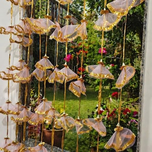 Könnte beinhalten: Dekorative Fensterdekoration mit zahlreichen kleinen, schirmförmigen Ornamenten. Die Ornamente bestehen aus durchsichtigem, hellrosa Stoff mit goldenem Besatz und sind an goldfarbenen Schnüren aufgehängt. Der Hintergrund zeigt eine Gartenszene.