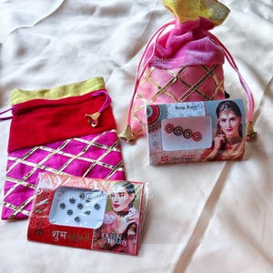 May include: Two pink drawstring bags with gold accents and a red bag with gold trim sit on a cream-colored surface. Two boxes of decorative face jewels are also present, one with the text "Roop Rajni" and the other with "Queen Hearts".