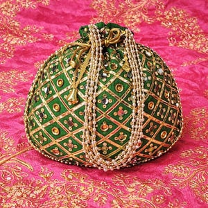 May include: A green, round, beaded pouch with a drawstring closure. The pouch is decorated with gold accents and a diamond pattern. A strand of white pearls hangs from the top. The pouch is set against a pink background.