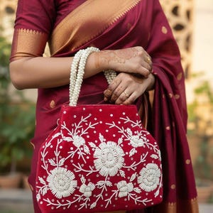 Peut inclure: Un sac à main en velours rouge avec des broderies florales blanches et une poignée de perles. Le sac est tenu contre un sari bordeaux avec une bordure dorée. Le design du sac présente de grandes fleurs stylisées.