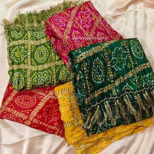 May include: A selection of vibrant patterned scarves in green, red, pink, and yellow. Each scarf displays a unique design with detailed patterns and fringe. The scarves are folded and arranged on a light-coloured fabric.