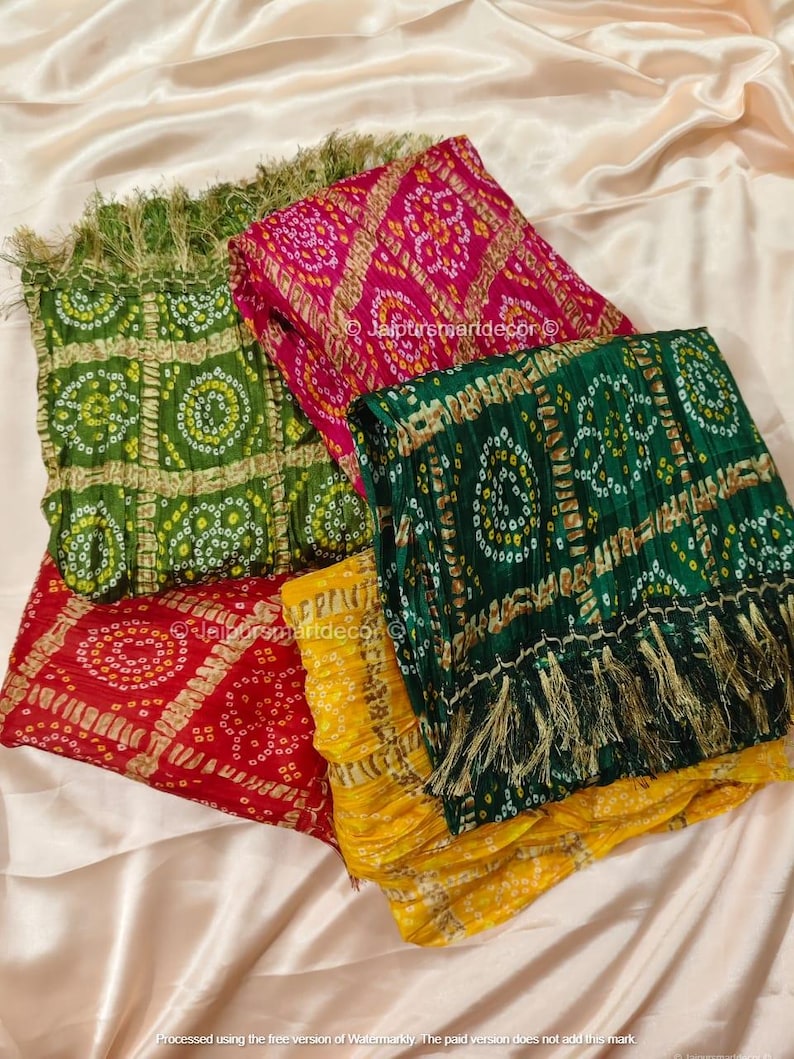 May include: A selection of vibrant patterned scarves in green, red, pink, and yellow. Each scarf displays a unique design with detailed patterns and fringe. The scarves are folded and arranged on a light-coloured fabric.