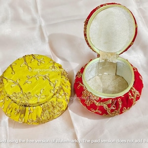 Könnte beinhalten: Zwei runde, dekorative Boxen. Eine ist gelb mit goldener Stickerei, die andere ist rot mit goldener Stickerei und ist geöffnet, wodurch ein weißes Innere sichtbar wird. Die Boxen befinden sich auf einer weißen Oberfläche.
