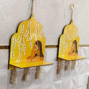 Può includere: Due mensole da parete in legno giallo con un design tradizionale indiano. Ogni mensola presenta un'immagine dipinta di una donna, dettagli floreali bianchi e un nappa di perline. Le mensole sono appese a un muro strutturato.