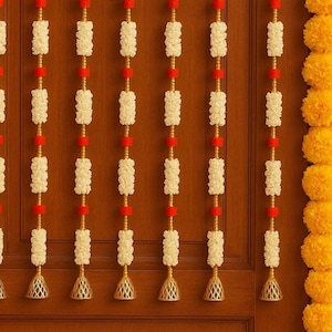 Puede incluir: Guirnalda decorativa para puerta con hileras de cuentas blancas texturizadas, detalles rojos y colgantes dorados en forma de campana. La guirnalda está colocada contra una puerta de madera marrón, con flores amarillas de caléndula a la derecha.