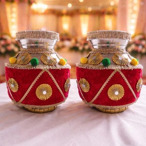 Peut inclure: Deux vases décoratifs rouges et dorés avec des dessus argentés. Les vases ont une texture velours avec des accents dorés, notamment des ornements circulaires et des bordures. Ils sont ornés de pompons jaunes et verts. L'arrière-plan est un cadre intérieur flou.