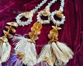 May include: Detailed shot of decorative garlands with silver and gold detailing. The garlands have silver loops, gold floral elements, and long tassels with colourful threads. The background is a deep burgundy velvet fabric.