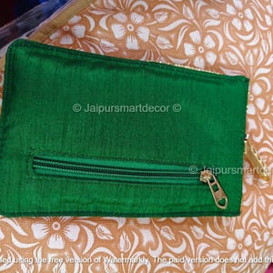 May include: A rectangular, emerald green pouch with a zipper closure and a gold-colored zipper pull. The pouch is made of a shiny fabric and is set against a floral patterned background. The text "Jaipursmartdecor" is visible.