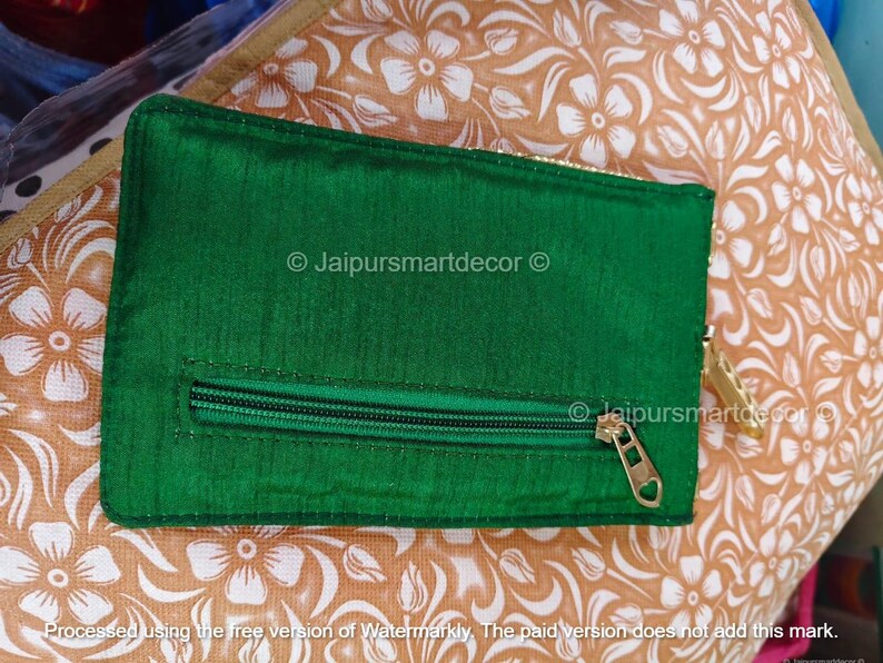 May include: A rectangular, emerald green pouch with a zipper closure and a gold-colored zipper pull. The pouch is made of a shiny fabric and is set against a floral patterned background. The text "Jaipursmartdecor" is visible.