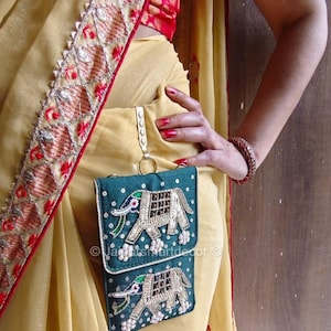 May include: Teal embroidered phone pouch with an elephant design. The pouch is adorned with silver and gold sequins and white pearl-like beads. The background is a light yellow saree with red and gold trim.
