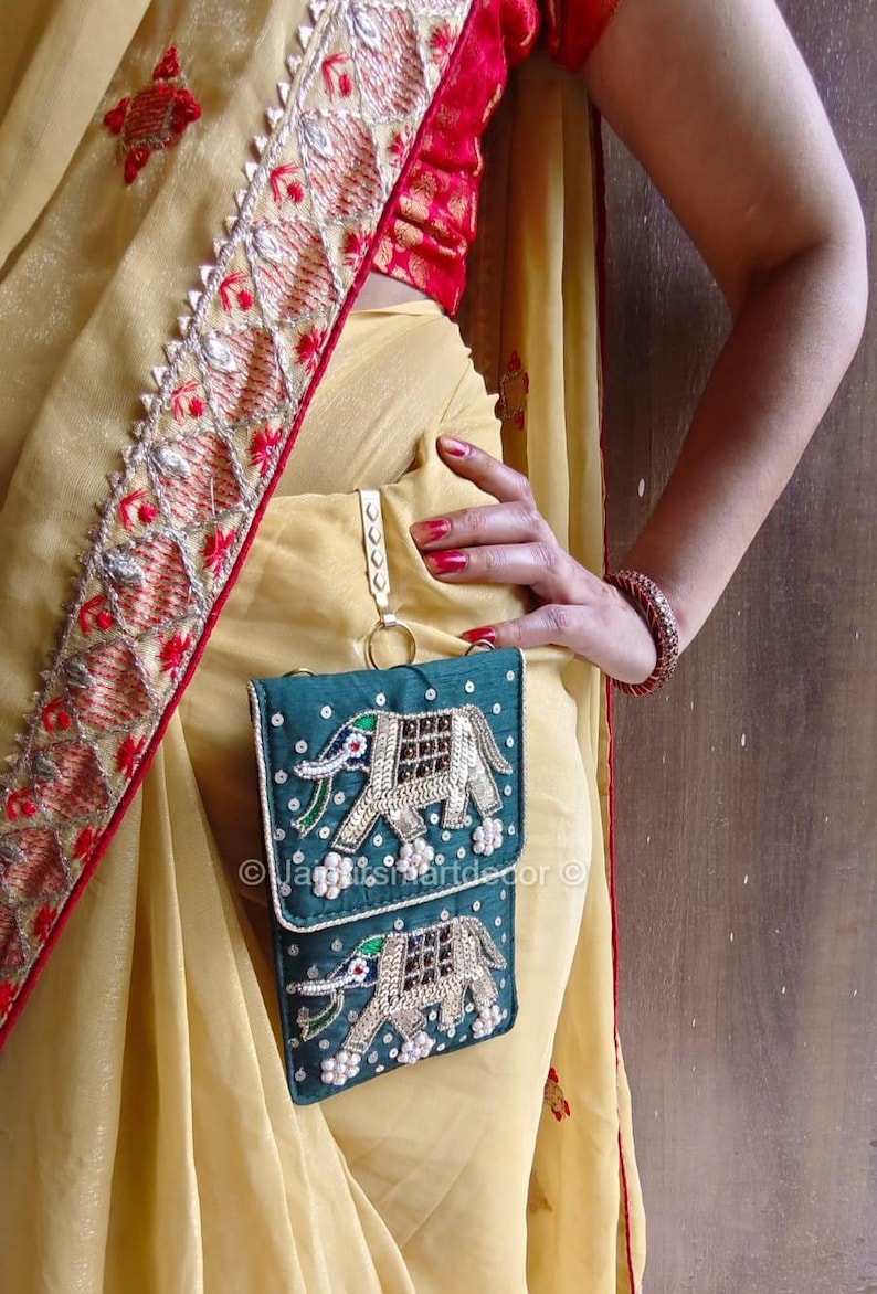 May include: Teal embroidered phone pouch with an elephant design. The pouch is adorned with silver and gold sequins and white pearl-like beads. The background is a light yellow saree with red and gold trim.