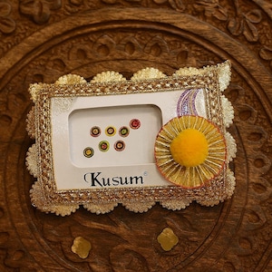 May include: A decorative gold-framed item with a white interior, featuring colorful dots and the brand name "Kusum". A yellow pom-pom and a decorative element are attached. The item rests on a carved wooden surface.