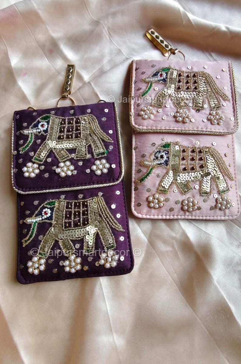 May include: Purple and pink embroidered elephant design phone pouches. The pouches feature a flap closure and a gold-colored chain. The elephant design is embellished with sequins and beads. The pouches are likely made of a satin-like material.