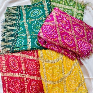 May include: A selection of vibrantly coloured scarves with fringed borders. The scarves display intricate circular patterns in green, pink, red, and yellow. The fabric has a soft, draped appearance with a slight sheen, suggesting a delicate texture.