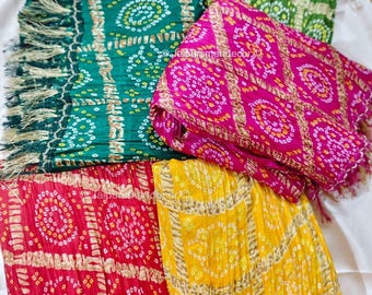 May include: A collection of colorful, patterned scarves with fringed edges. The scarves are in shades of green, pink, red, and yellow, each featuring a detailed, circular design. The fabric appears soft and draped, with a subtle sheen.