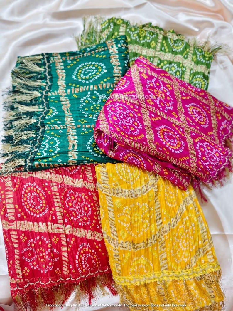 May include: A selection of vibrantly coloured scarves with fringed borders. The scarves display intricate circular patterns in green, pink, red, and yellow. The fabric has a soft, draped appearance with a slight sheen, suggesting a delicate texture.