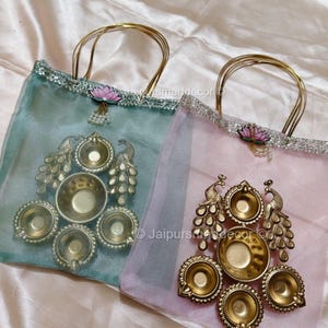 May include: Two decorative bags with gold handles. One is light blue, the other is pink. Each bag features a gold peacock design and a set of small bowls. The bags are adorned with a lotus flower and pearl accents.