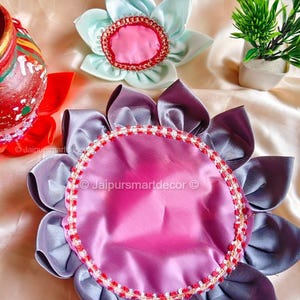 May include: Decorative floral-shaped items in shades of pink, lavender, and light blue. The petals are made of satin-like fabric, and the centers are adorned with a contrasting trim. The items are arranged on a satin surface.