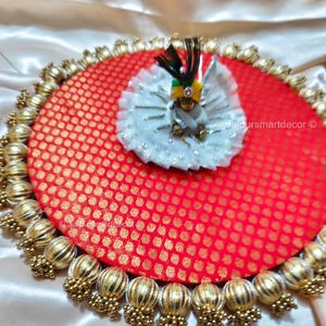 May include: A decorative, round red and gold fabric base with a small doll wearing a white dress and a feathered headdress. The base is trimmed with gold-coloured beads and tassels.