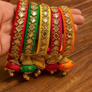 May include: A collection of colorful bangles in red, green, yellow, pink, and orange. Each bangle is adorned with square mirrored accents and gold-colored embellishments. The bangles have a bell-shaped charm at the bottom.