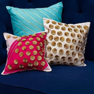 May include: Three decorative throw pillows in shades of pink, white, and turquoise. The pink pillow features gold circular embellishments and a diagonal design. The white pillow is covered in gold circular accents, and the turquoise pillow has gold stripes.