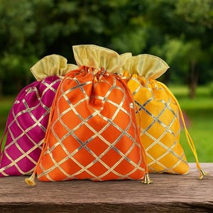 May include: Three colorful drawstring bags in magenta, orange, and yellow, each with a gold lattice pattern. The bags have gold accents and are set on a wooden surface against a blurred green background.