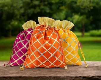May include: Three colorful drawstring bags in magenta, orange, and yellow, each with a gold lattice pattern. The bags have gold accents and are set on a wooden surface against a blurred green background.