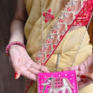 May include: A bright pink, embroidered crossbody bag with a silver chain strap. The bag features an elephant design with silver sequins and green and red accents. The person is wearing a gold-colored sari with red trim and a matching bracelet.