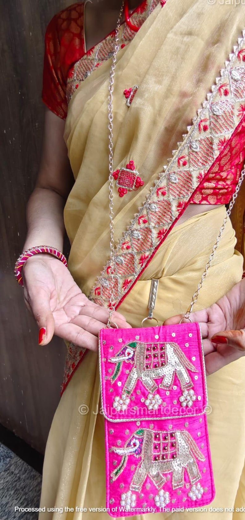 May include: A bright pink, embroidered crossbody bag with a silver chain strap. The bag features an elephant design with silver sequins and green and red accents. The person is wearing a gold-colored sari with red trim and a matching bracelet.
