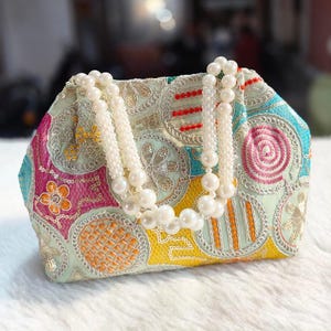 Peut inclure: Un sac à main brodé aux couleurs vives, orné d'une poignée de perles. Le sac présente des motifs floraux et géométriques dans des tons de rose, jaune, bleu et or. La poignée est faite de perles blanches.