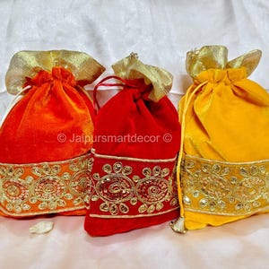 Op de afbeelding: Drie fluwelen trekkoordzakjes in oranje, rood en geel. Elk zakje heeft een goudkleurige, decoratieve rand met pailletten en een bijpassende gouden trekkoordsluiting. De zakjes worden waarschijnlijk gebruikt voor cadeaus of kleine items.