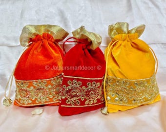 May include: Three velvet drawstring bags in orange, red, and yellow. Each bag has a gold-colored, decorative trim with sequins and a matching gold drawstring closure. The bags are likely used for gifts or small items.