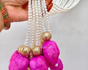 May include: A strand of white pearl beads with pink fabric flower accents and gold-colored spherical embellishments. The decorative piece is held against a white background.