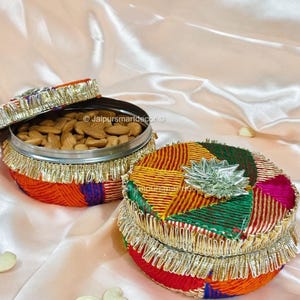 May include: Two round, colorful, decorative storage containers with lids. One container is open, revealing almonds. The containers are adorned with gold fringe and a silver embellishment. The containers are on a satin surface.