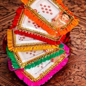 Könnte beinhalten: Ein Stapel bunter, dekorativer Tabletts mit weißen Zentren und leuchtenden Verzierungen. Jedes Tablett hat eine andere Farbe: Orange, Rot, Gelb, Grün und Pink. Goldene Verzierungen und ein Bild einer Frau schmücken die Tabletts.
