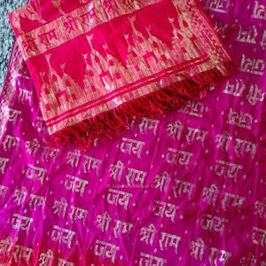 May include: A bright pink and yellow textile with silver text and a matching scarf. The fabric features repeating text and a decorative border on the scarf. The scarf has a fringe.