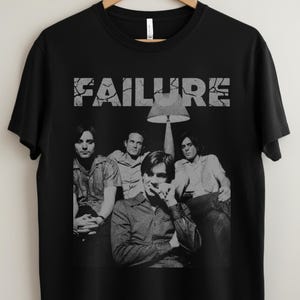 Op de afbeelding: Zwart t-shirt met een grijswaardenafbeelding van de band Failure, met de naam van de band in een vervormd lettertype boven de afbeelding. De afbeelding toont de bandleden zittend onder een lamp.
