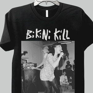 Op de afbeelding: Zwart T-shirt met de witte tekst "B:KN: KILL". De afbeelding toont een zwart-wit foto van een band die optreedt. Het T-shirt hangt aan een zilveren kledinghanger.