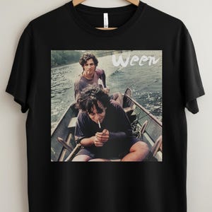 May include: Black t-shirt featuring a vintage image of two people in a boat, with the band name "Ween" in white text. The image shows the people lighting cigarettes. The background shows water and trees.