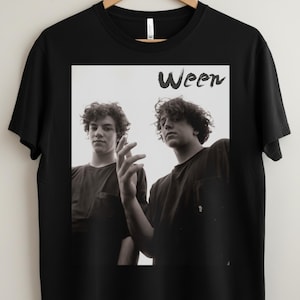 Op de afbeelding: Zwart T-shirt met een zwart-wit afbeelding van twee mensen met krullend haar. Het woord "Ween" is gedrukt in een gestileerd lettertype boven de afbeelding. Het T-shirt heeft een ronde hals en korte mouwen.