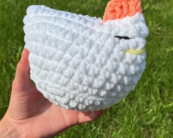 May include: A handmade white crochet chicken with a peach-colored comb and a yellow beak. The chicken has a black eye and is made of soft, textured yarn. The craft item is held in a hand against a green grass background.