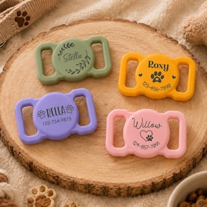 May include: Four silicone pet collar tags in sage green, mustard yellow, lavender, and light pink. Each rectangular tag features a central circular area with custom names, paw prints, and phone numbers printed in black, designed to slide onto a collar.