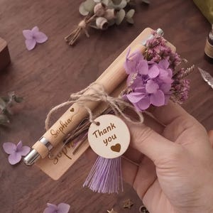 Puede incluir: Un bolígrafo de madera clara grabado con el nombre Sophia, atado con cordel a un estuche de madera a juego. El conjunto está decorado con flores secas moradas, una pequeña etiqueta redonda que dice Thank you con un corazón y una borla morada.