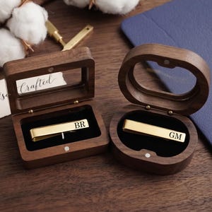 May include: Two personalized gold-colored tie clips, each in a wooden box. One clip is engraved with "BR", the other with "GM". The boxes are open, revealing the clips against a black velvet lining. The word "Crafted" is visible.