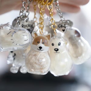 Puede incluir: Una colección de llaveros de resina transparente con diseños de animales, incluyendo un gato, un corgi y un perro. Cada llavero tiene una cadena plateada o dorada y una pequeña etiqueta en forma de corazón con un nombre.