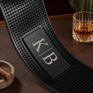 May include: A rectangular black rubber bar mat featuring a textured surface with raised circular nubs and a smooth central panel displaying the white capital letters K and B separated by a vertical line. The mat is flexible and durable.