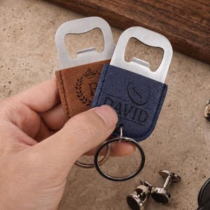 May include: Two metal bottle openers with leather-like covers in tan and navy blue. The tan cover features a crown and laurel wreath design, while the navy blue cover is embossed with the name DAVID and a coffee bean icon, each with a metal ring.